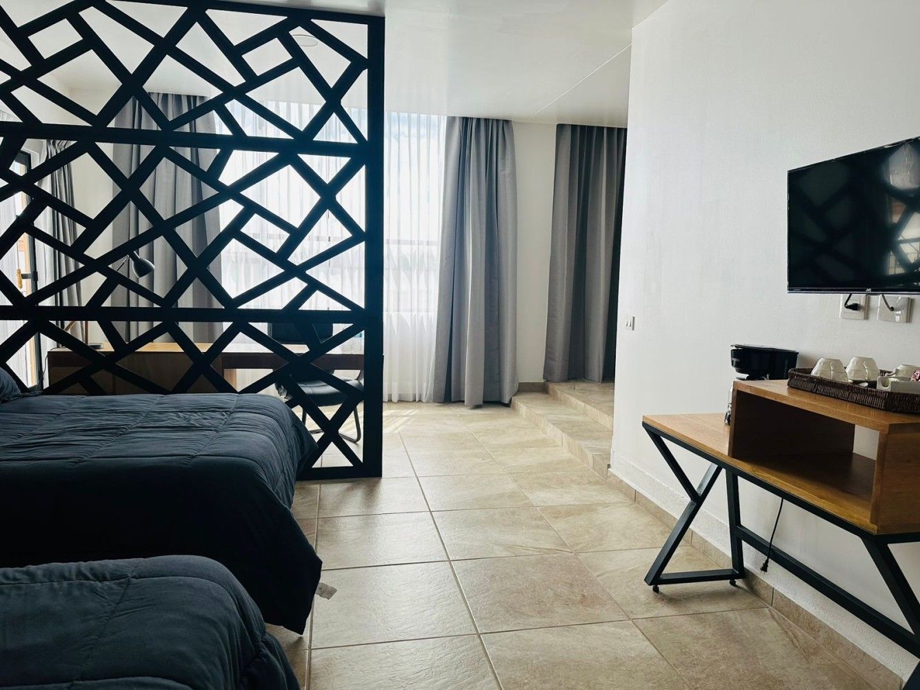 Interior de una habitación de hotel con separador de celosía negra, cama, televisor montado en la pared y escritorio plegable cerca de las cortinas.