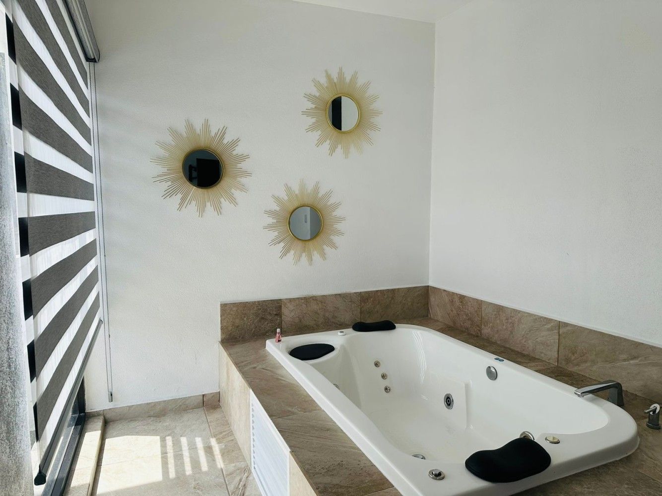 Baño con bañera de hidromasaje integrada, revestimiento de azulejos de piedra y persiana a rayas con decoración de pared en forma de rayos de sol.