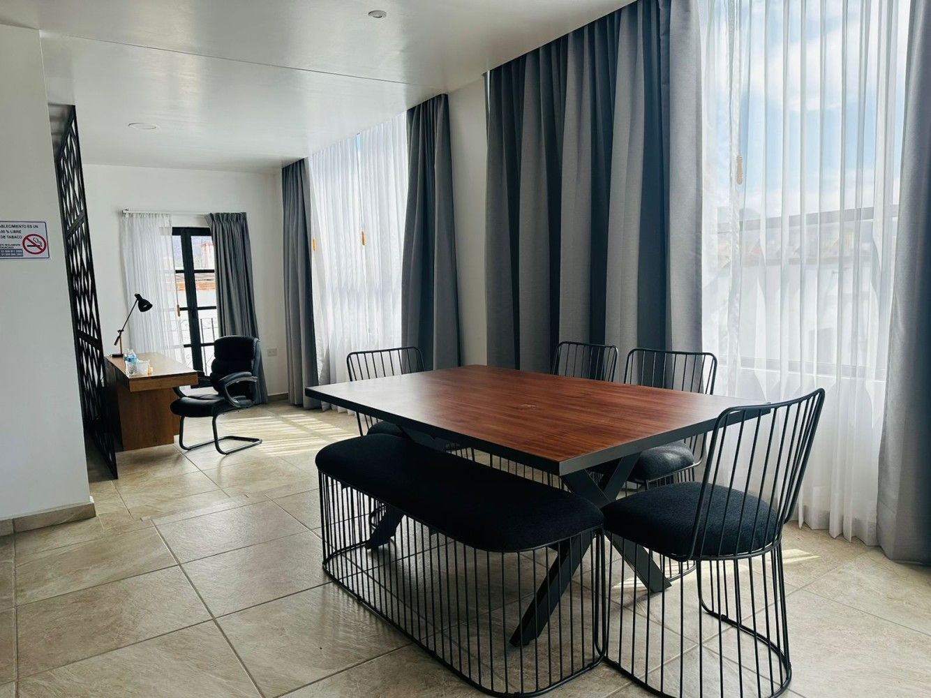 Comedor moderno con mesa de madera, sillas negras y cortinas que van del suelo al techo junto a grandes ventanales.