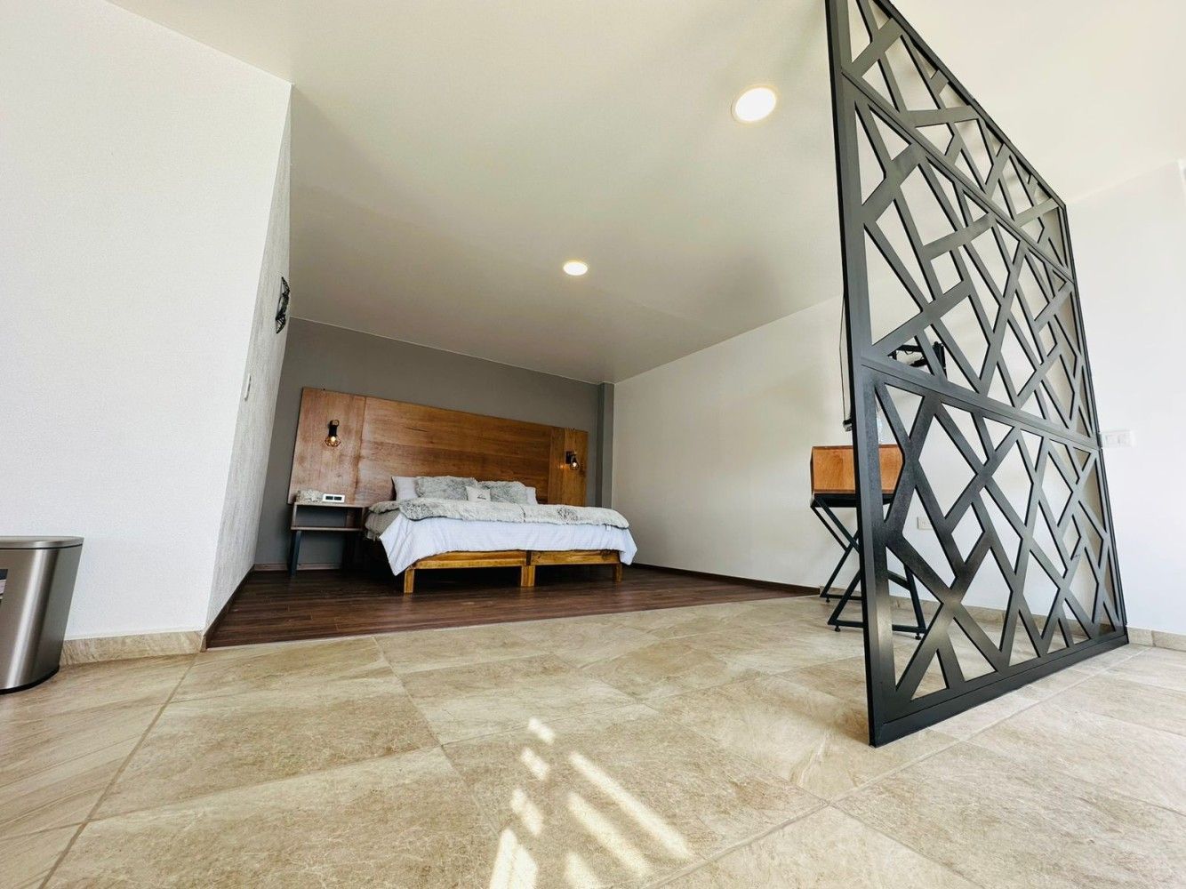 Dormitorio moderno con cama, cabecero de madera y separador de ambientes geométrico cerca de una puerta abierta.