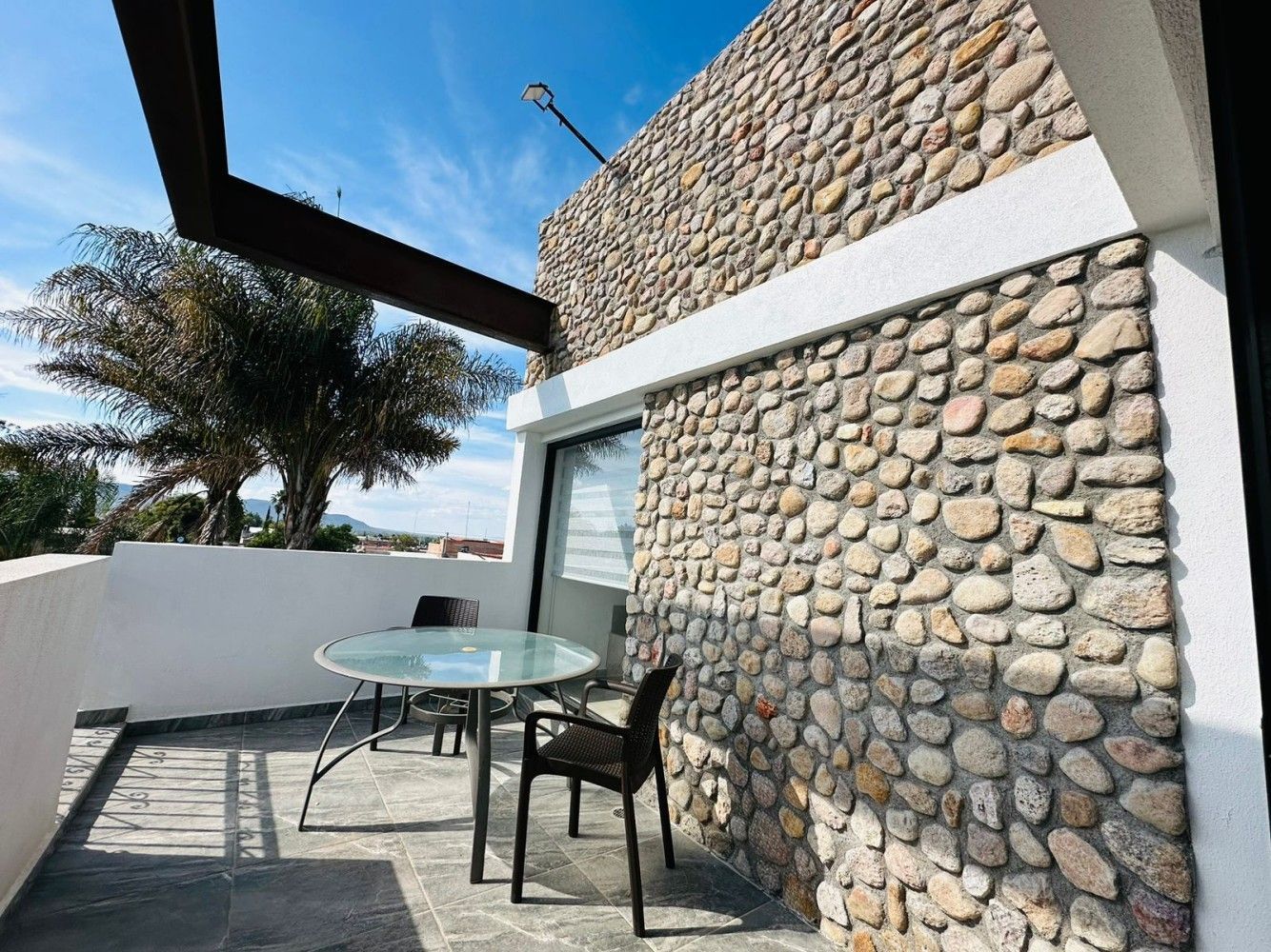 Patio con mesa y sillas de cristal junto a un muro de piedra y una gran ventana bajo el cielo azul.
