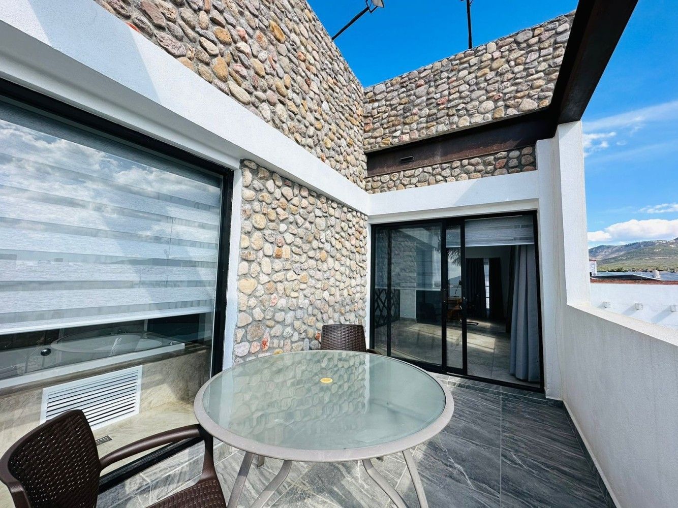 Terraza de piedra con mesa de cristal, puertas correderas y muros de piedra bajo un cielo azul.