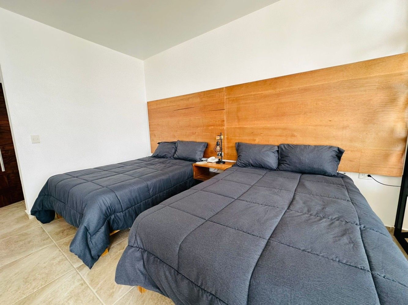 Dos camas bien hechas con edredones grises en un dormitorio luminoso y moderno con cabecero de madera.