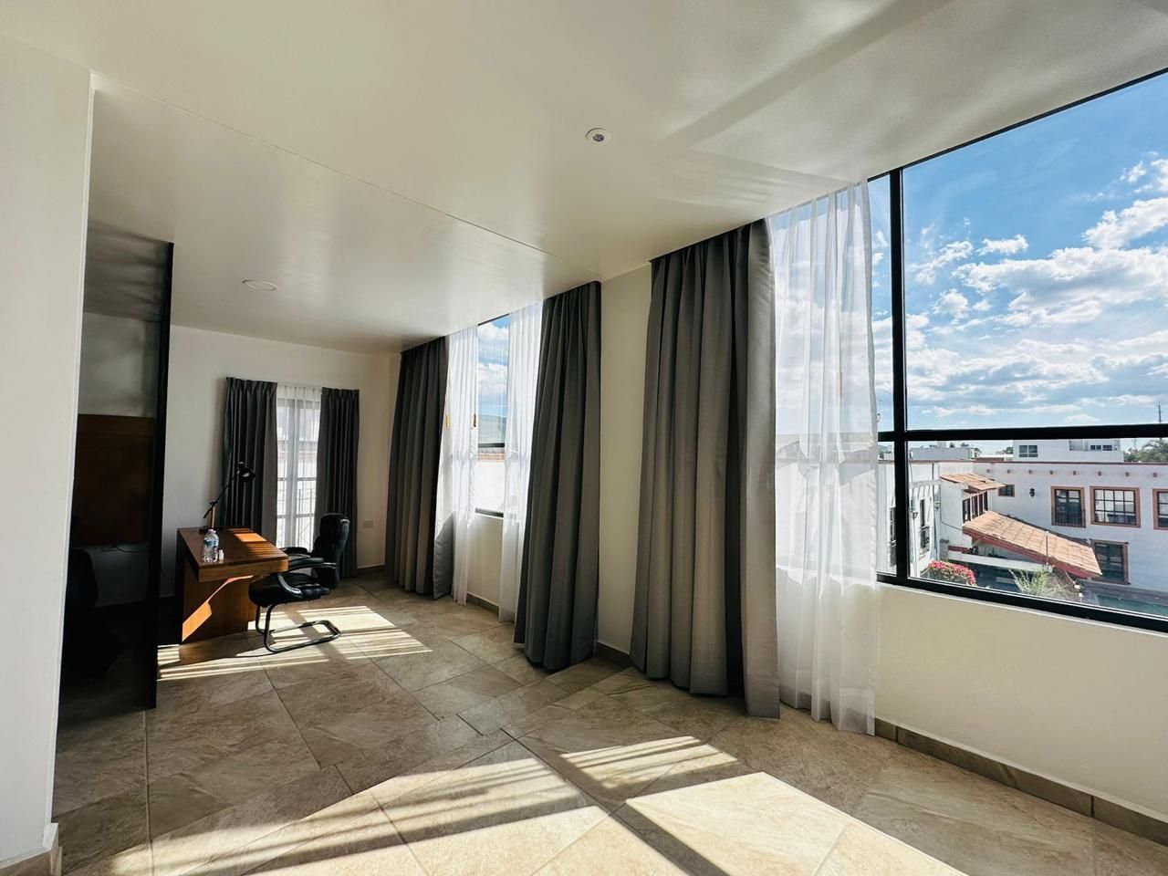 Habitación luminosa y vacía con grandes ventanales, cortinas grises y luz solar que proyecta sombras en el suelo.