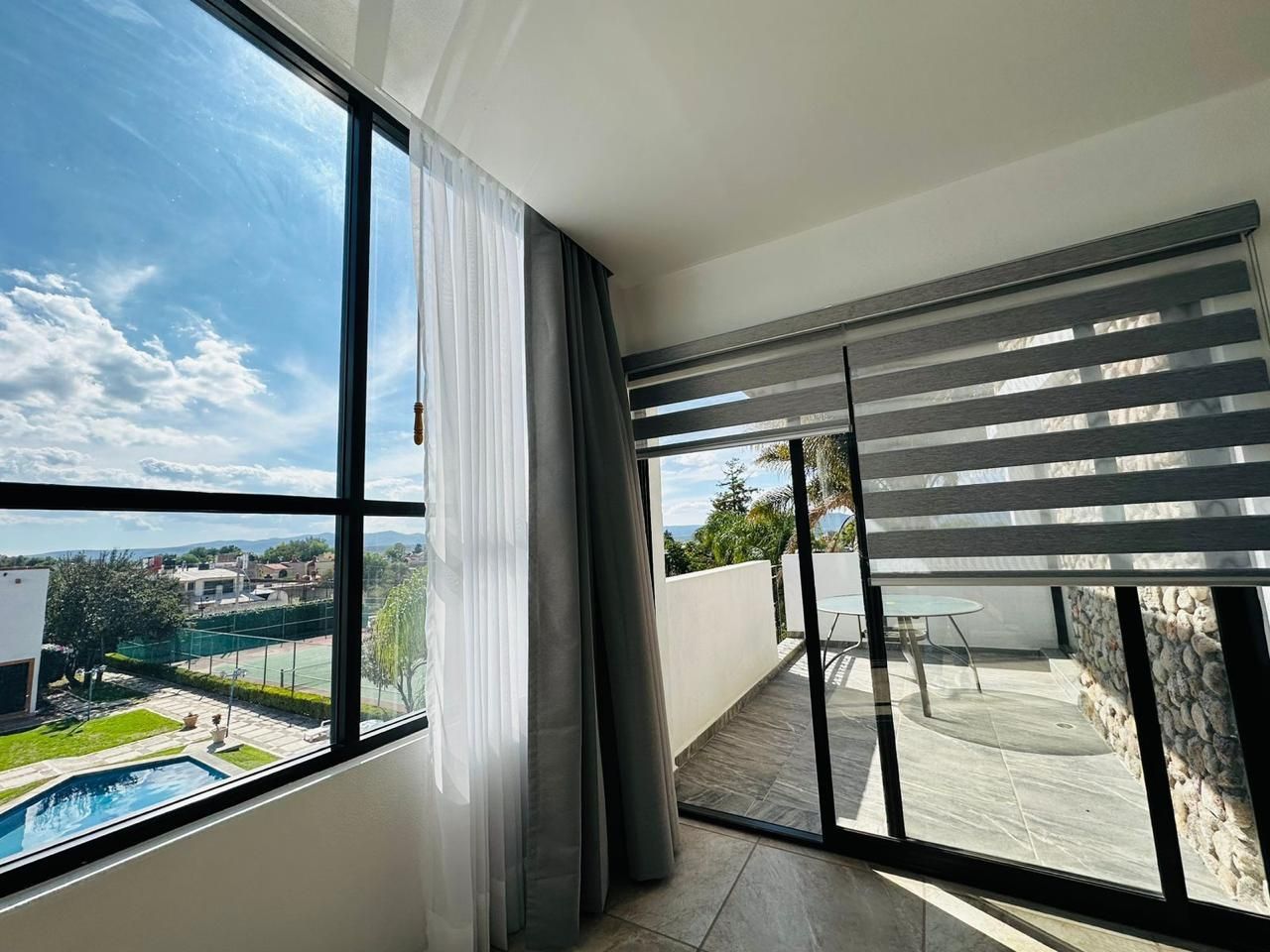 Dormitorio moderno con ventanales del suelo al techo, cortinas transparentes y puertas que dan a un balcón con vistas a la piscina y a la ciudad.
