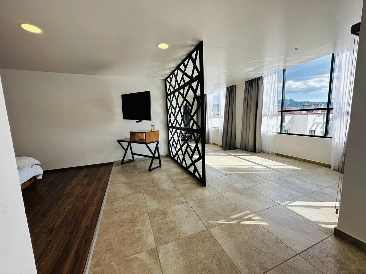 Habitación amplia y moderna con suelo de baldosas, cama, televisor, mesa plegable y grandes ventanales con cortinas.