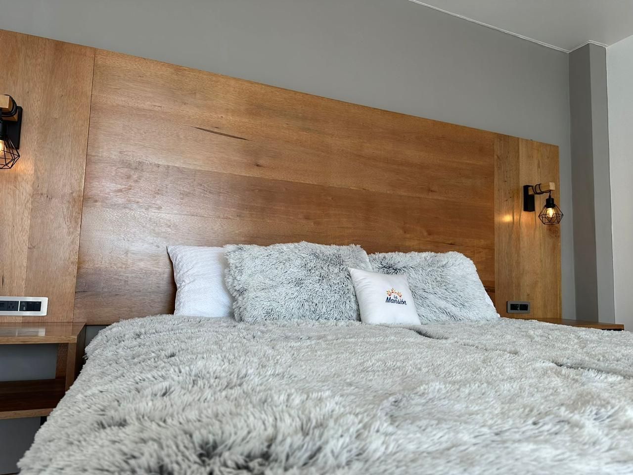 Dormitorio moderno con una cama grande, cabecero de madera, ropa de cama gris y apliques de pared.