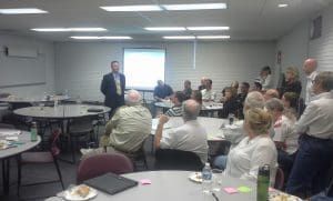 A man in a suit giving a presentation in a room of people seated at round tables.