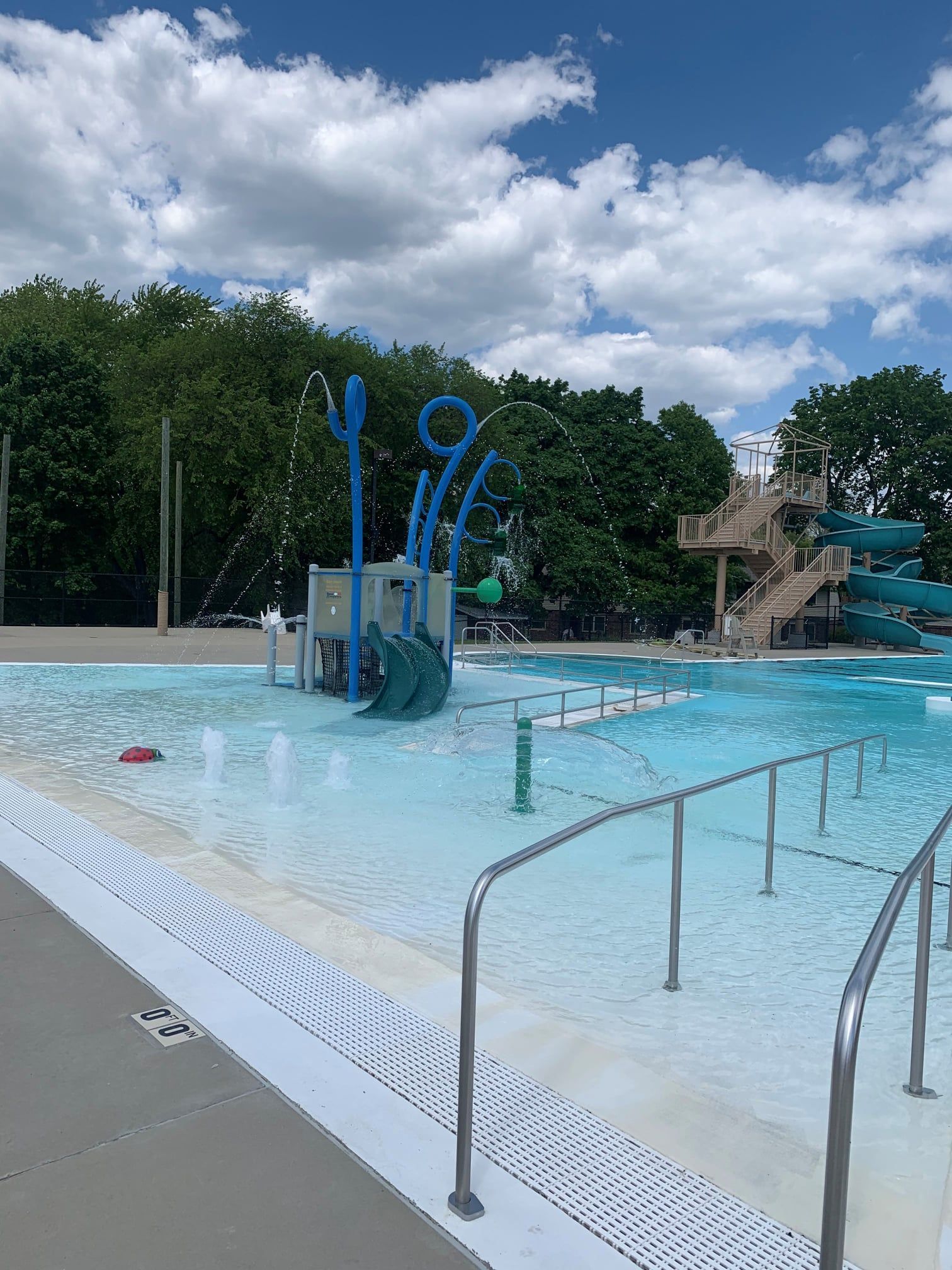 Outdoor swimming pool with blue splash features, metal handrails, and a waterslide under a cloudy sky