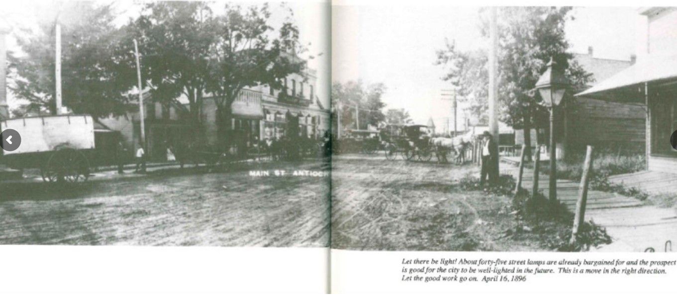Black and white photo of a dirt road with buildings and horse-drawn carriages.