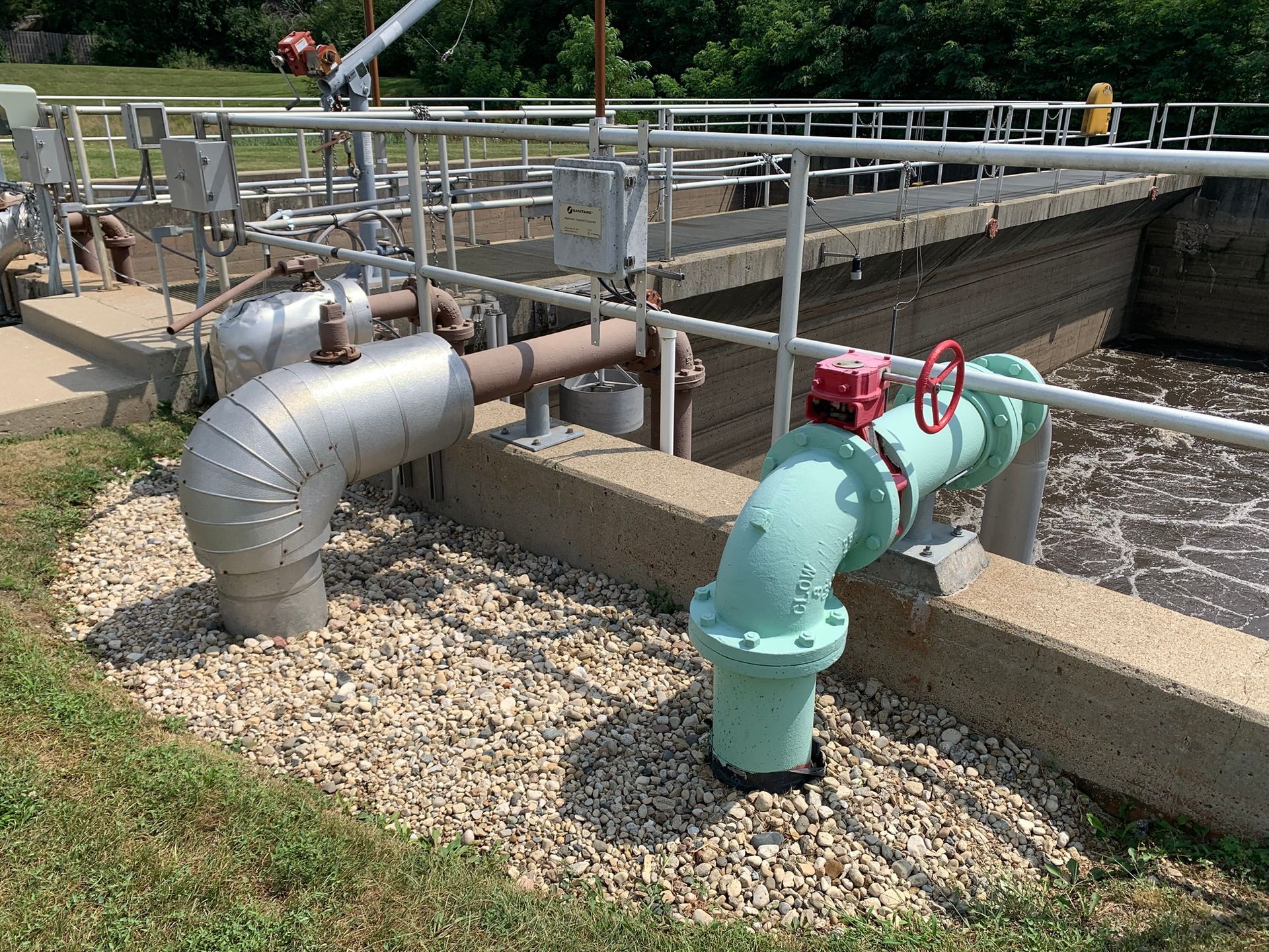 Pipes and a valve at a water treatment plant, with foamy water in the concrete basins, outdoors on a sunny day.
