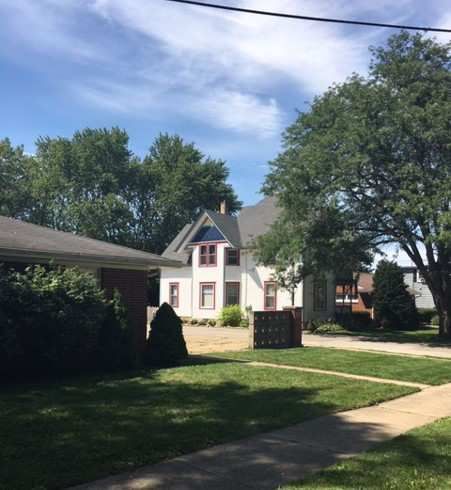 White house with dark roof and red trim, trees, lawn, and sidewalk on a sunny day.