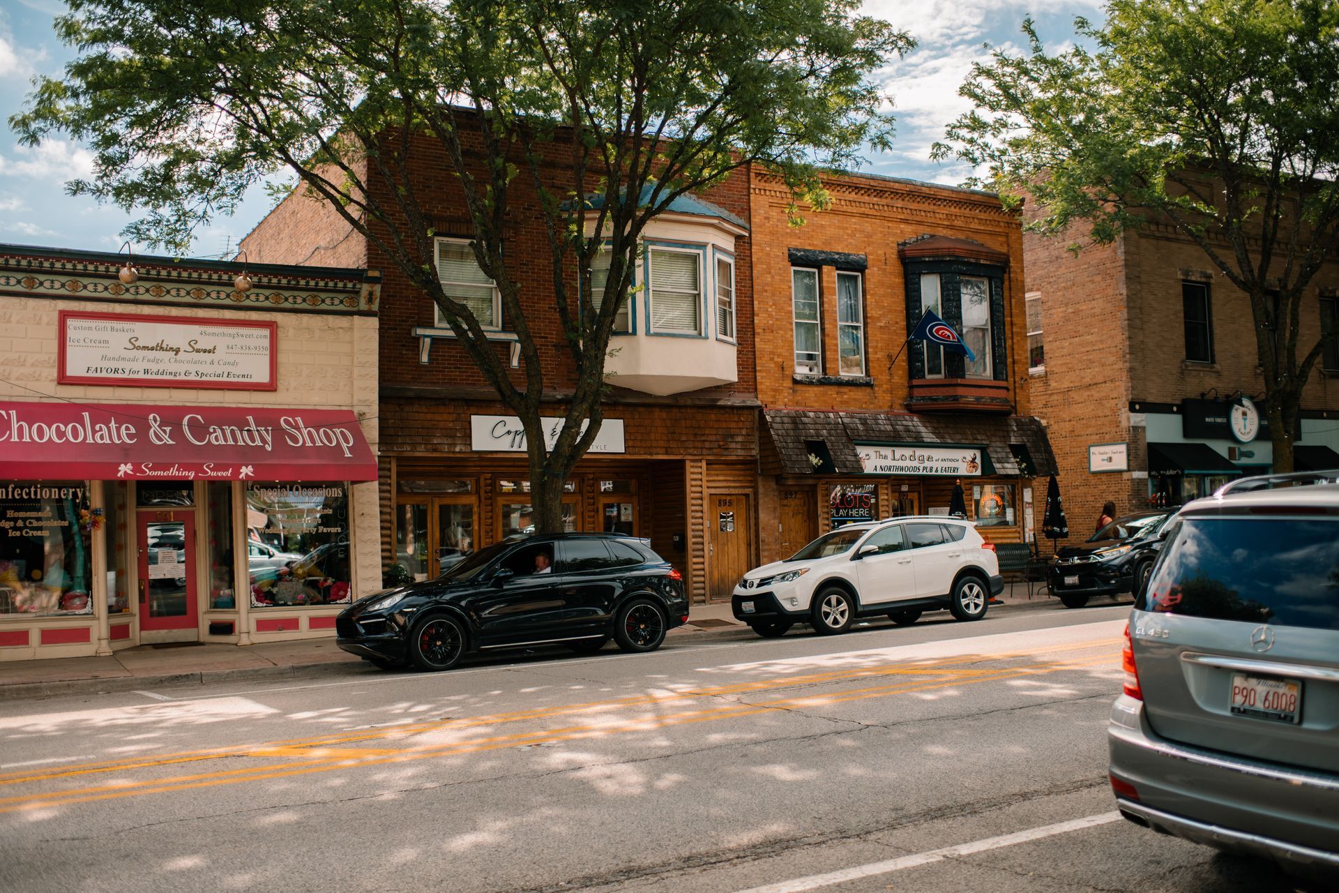 Street scene with parked cars and shops, including a chocolate shop with a pink awning.