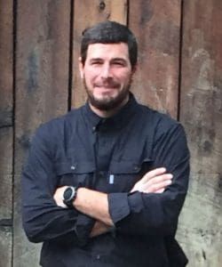 Man with beard, crossed arms, in front of a wooden wall, wearing a black shirt and watch.