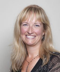Blonde woman with bangs smiles, wearing a black top and necklace, neutral background.