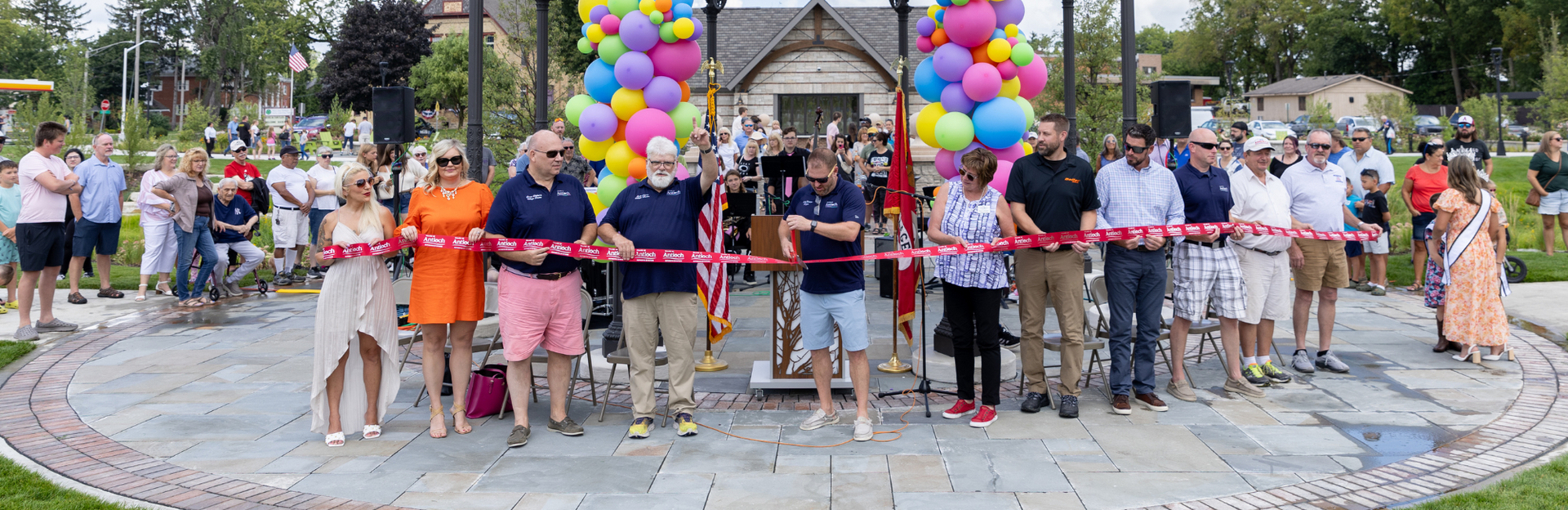 Group of people at a ribbon-cutting ceremony, many holding the ribbon in front of building with colorful balloons.