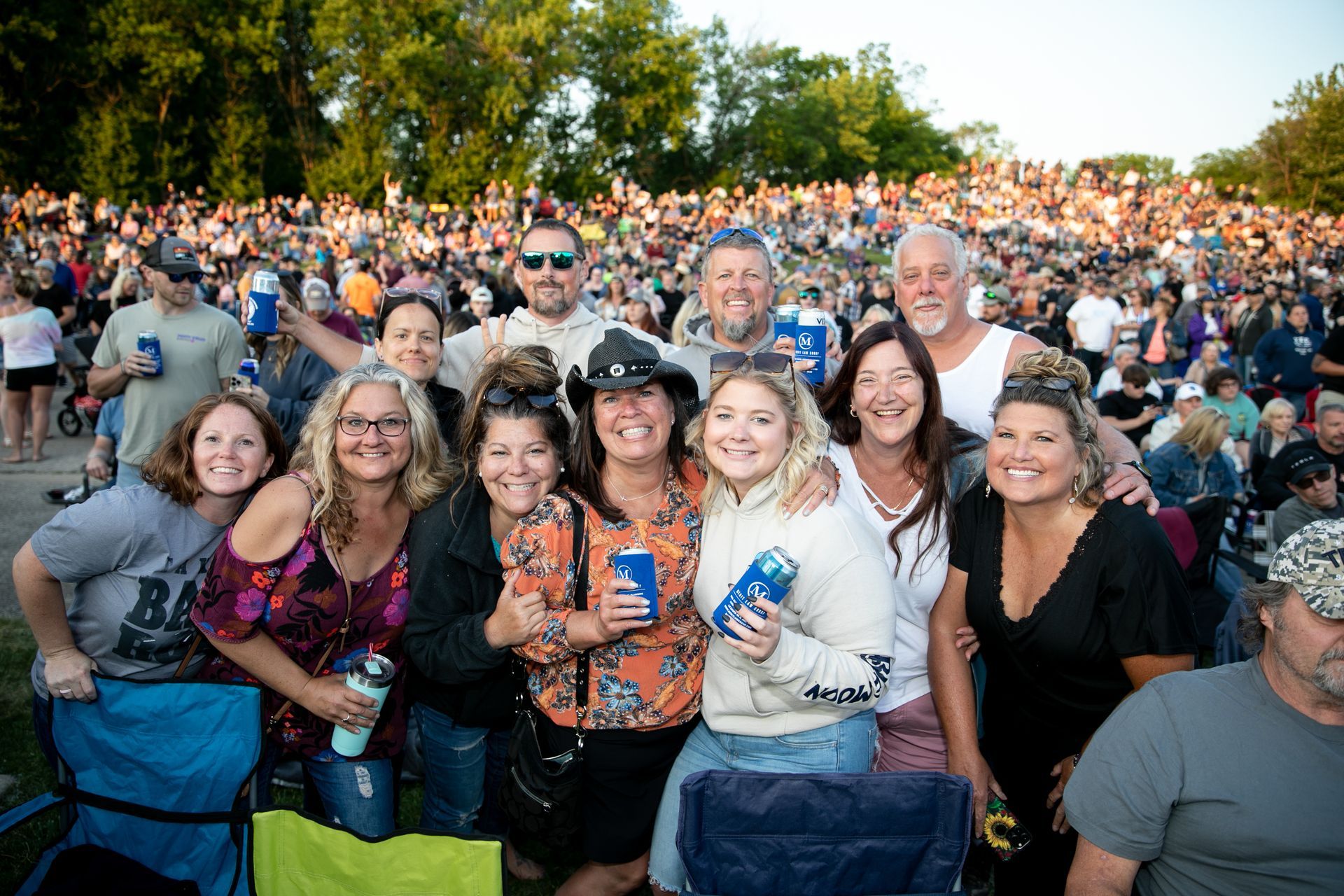 Group smiles at outdoor concert; crowd in background. People holding drinks, sunny setting.