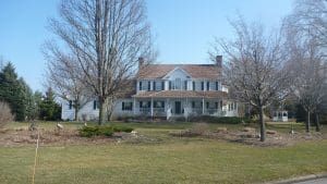 White two-story house with porch, black shutters, and bare trees in front yard on a sunny day.