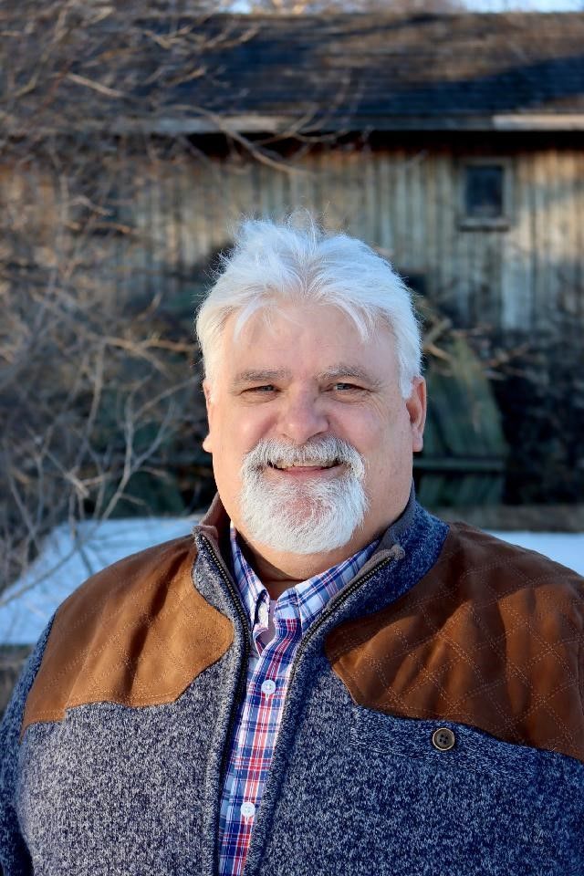 Man with white hair and beard smiling in front of a wooden building, wearing a blue sweater with brown shoulders.