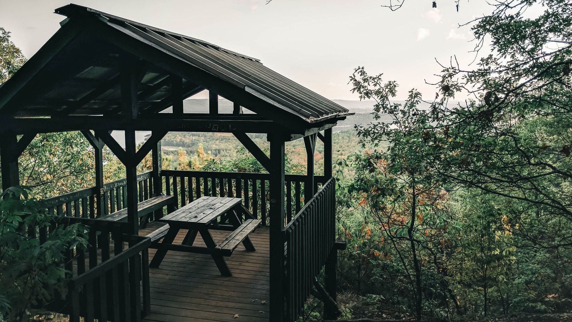Wooden gazebo with picnic table, surrounded by trees, overlooking a distant landscape.