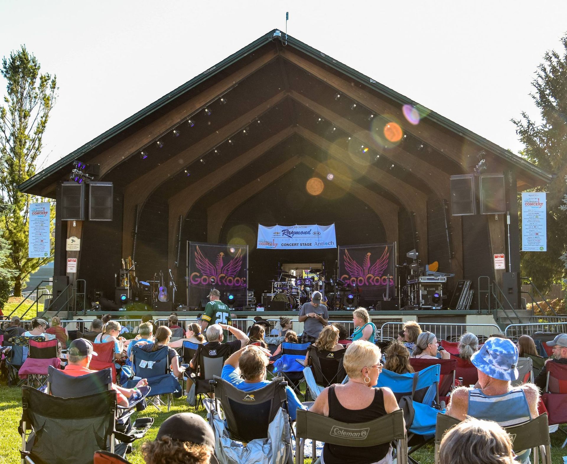 Outdoor concert at a wooden stage with an audience seated in folding chairs on the grass.