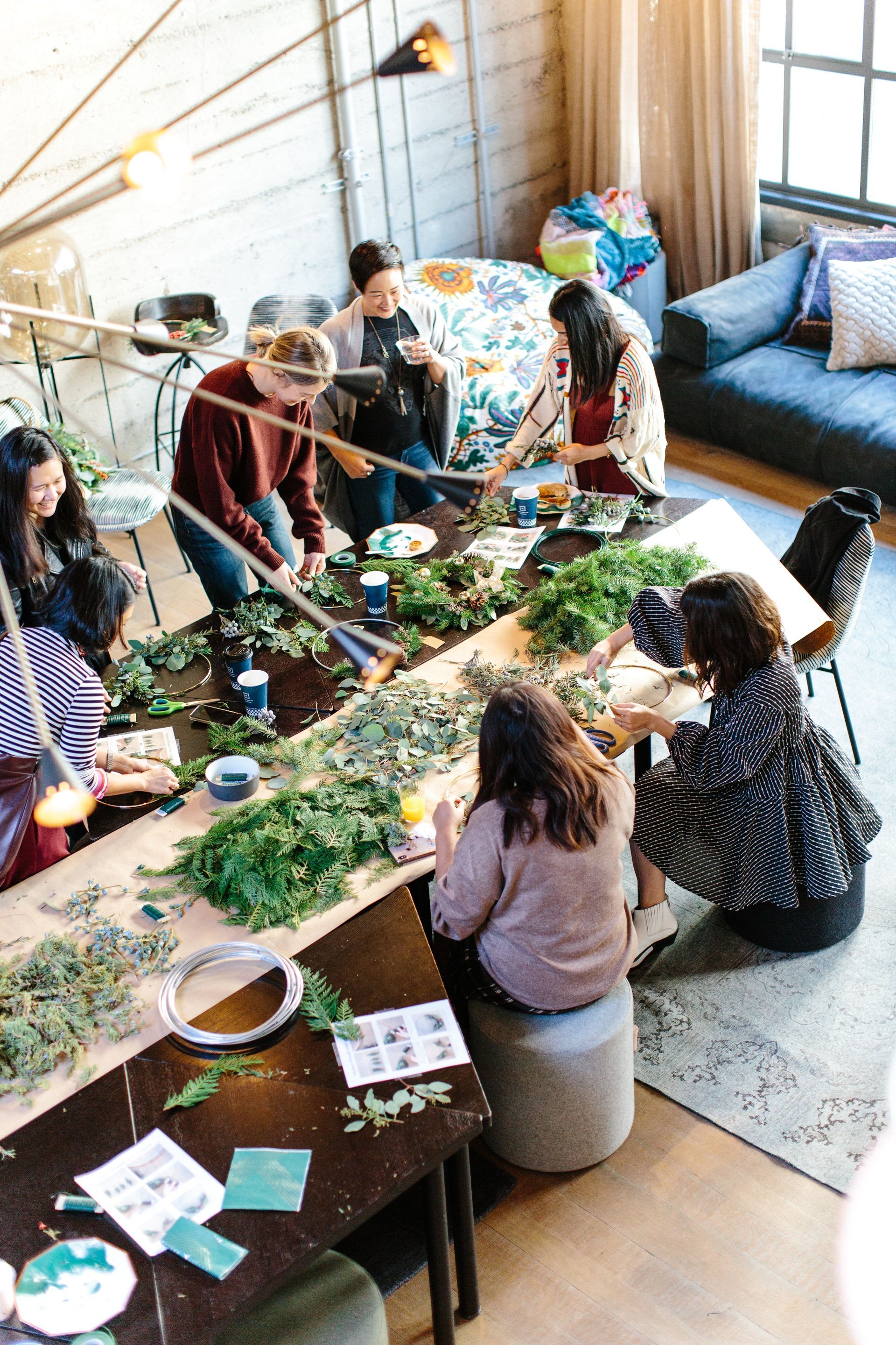 Group of women crafting greenery wreaths at a long table in a well-lit workshop space.