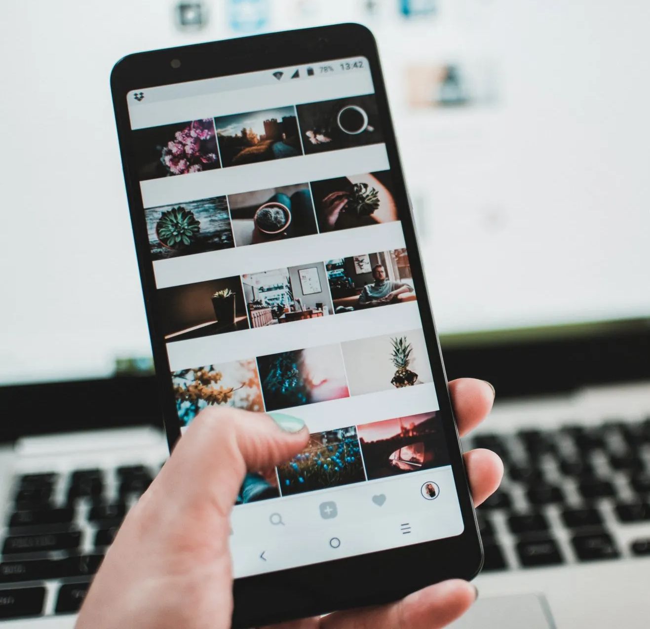 Person holding a black smartphone displaying a social media feed. A laptop is visible in the background.