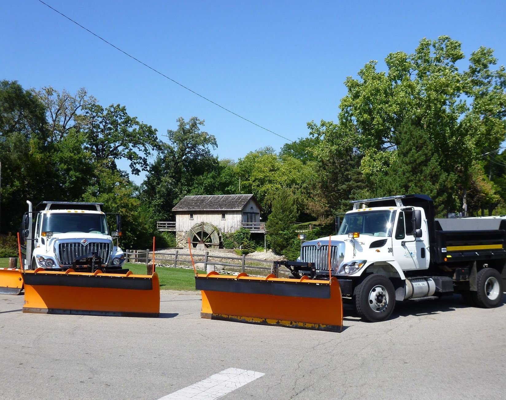 Two white snowplow trucks parked on asphalt in front of a wooden mill. Orange plows, trees, and blue sky.