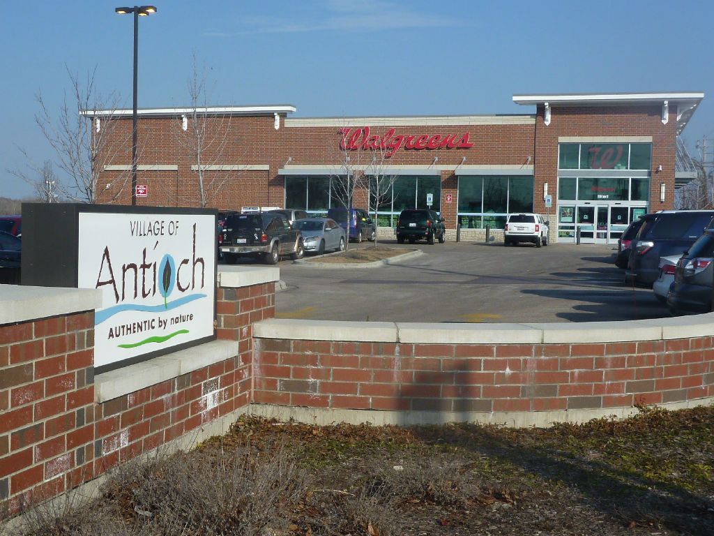Walgreens store in Antioch, Illinois, with a sign in front. Cars parked nearby, under a blue sky.