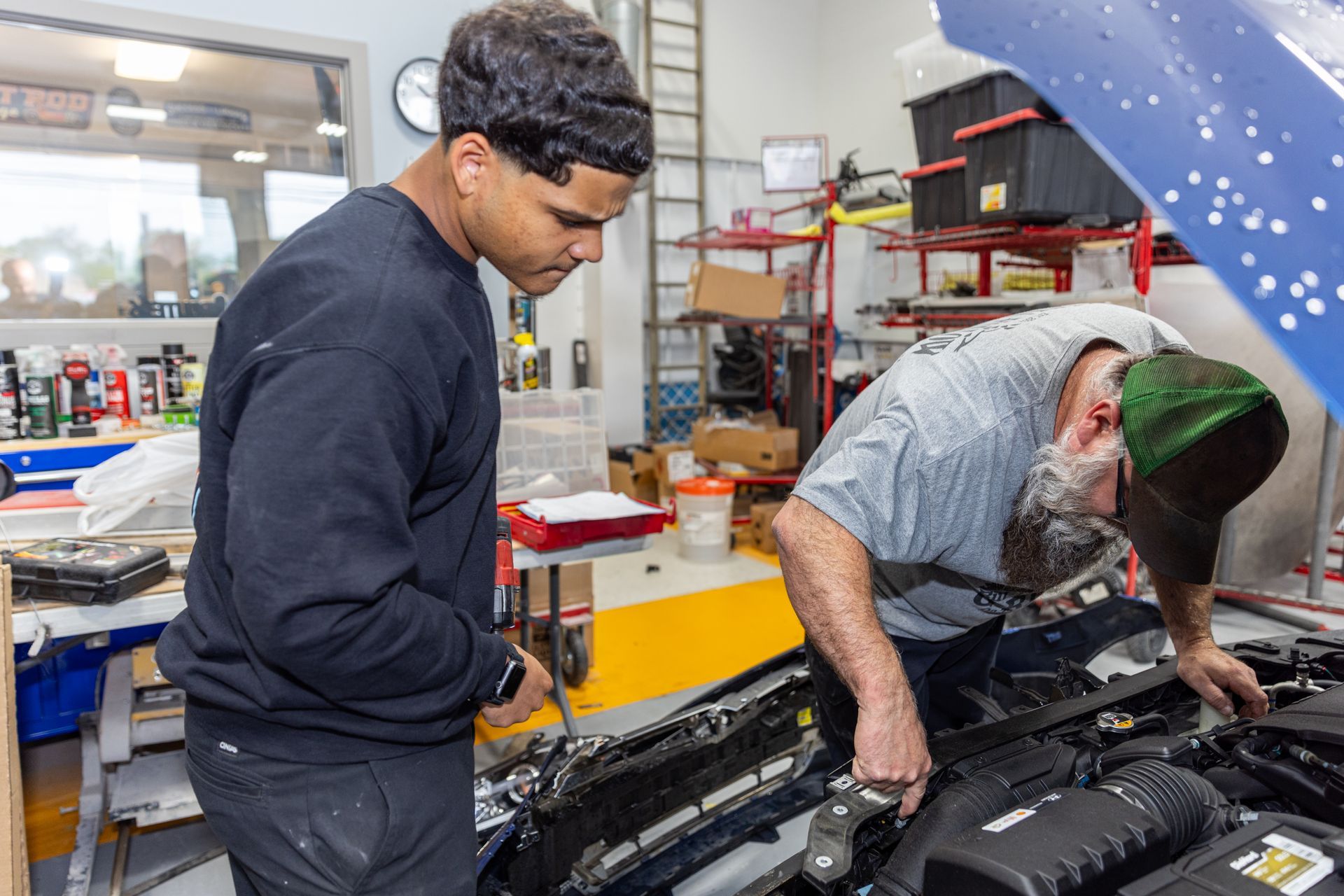 Two mechanics working on a car engine in a garage; one looks on, the other leans in close with a tool.