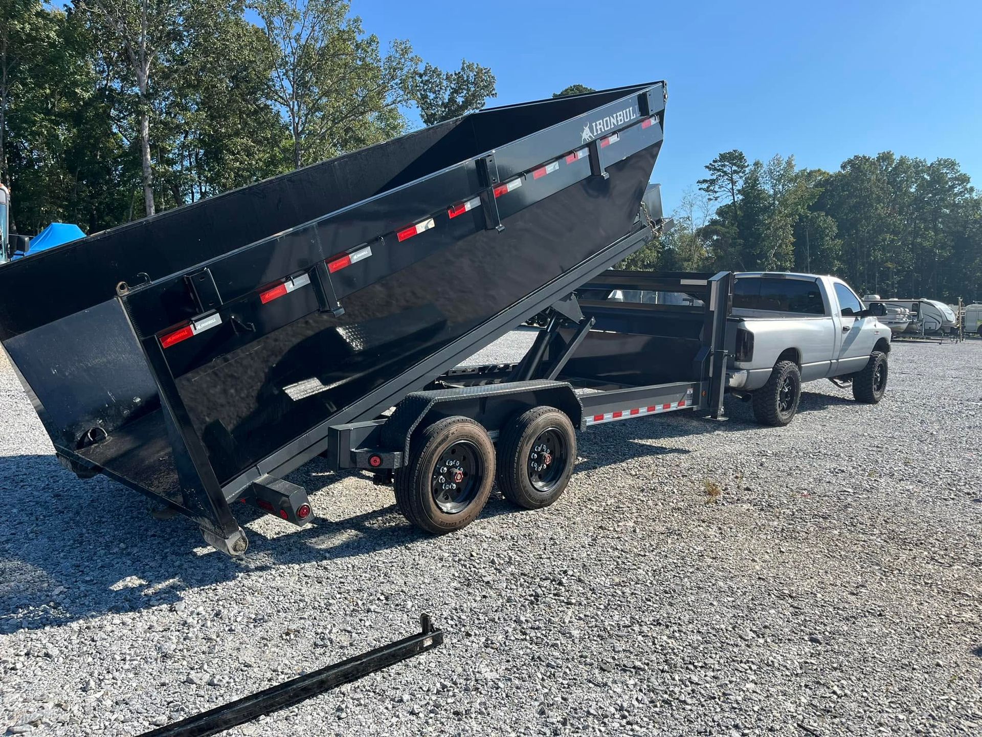 Black dump trailer lifted beside a silver pickup truck on a gravel lot under a clear blue sky