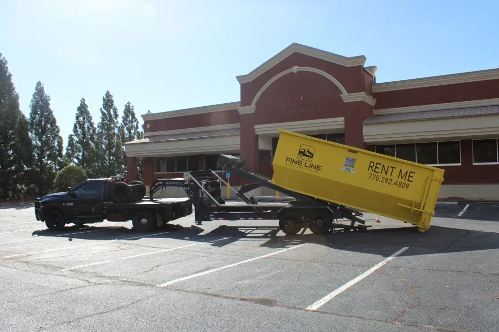 Tow truck lifting a yellow dumpster beside a brick building in a parking lot