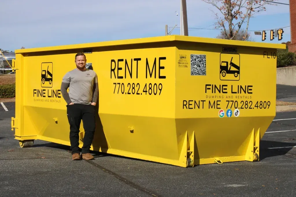 Man standing beside a yellow Fine Line rental dumpster with “RENT ME” signage on a parking lot