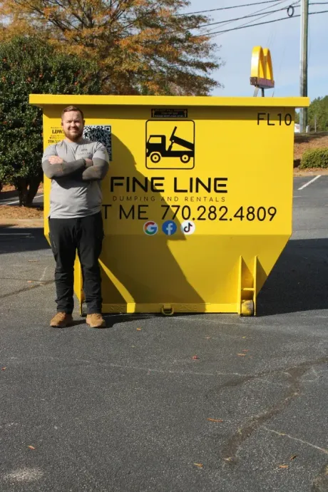 Man standing beside a bright yellow Fine Line dumpster in a parking lot on a sunny day