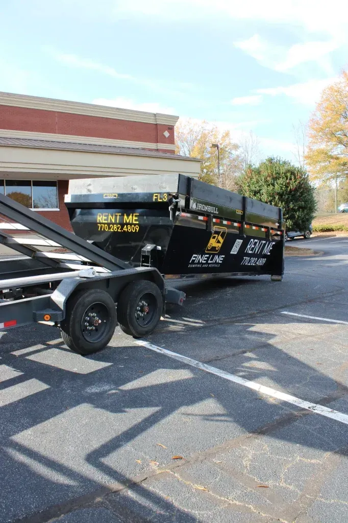 Black trailer with a dumpster parked in a striped parking lot beside a brick building and trees