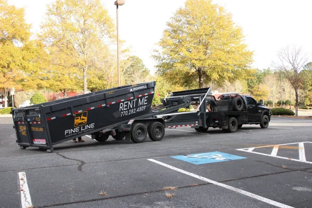 Tow truck hauling a black pickup and trailer in a parking lot with autumn trees nearby