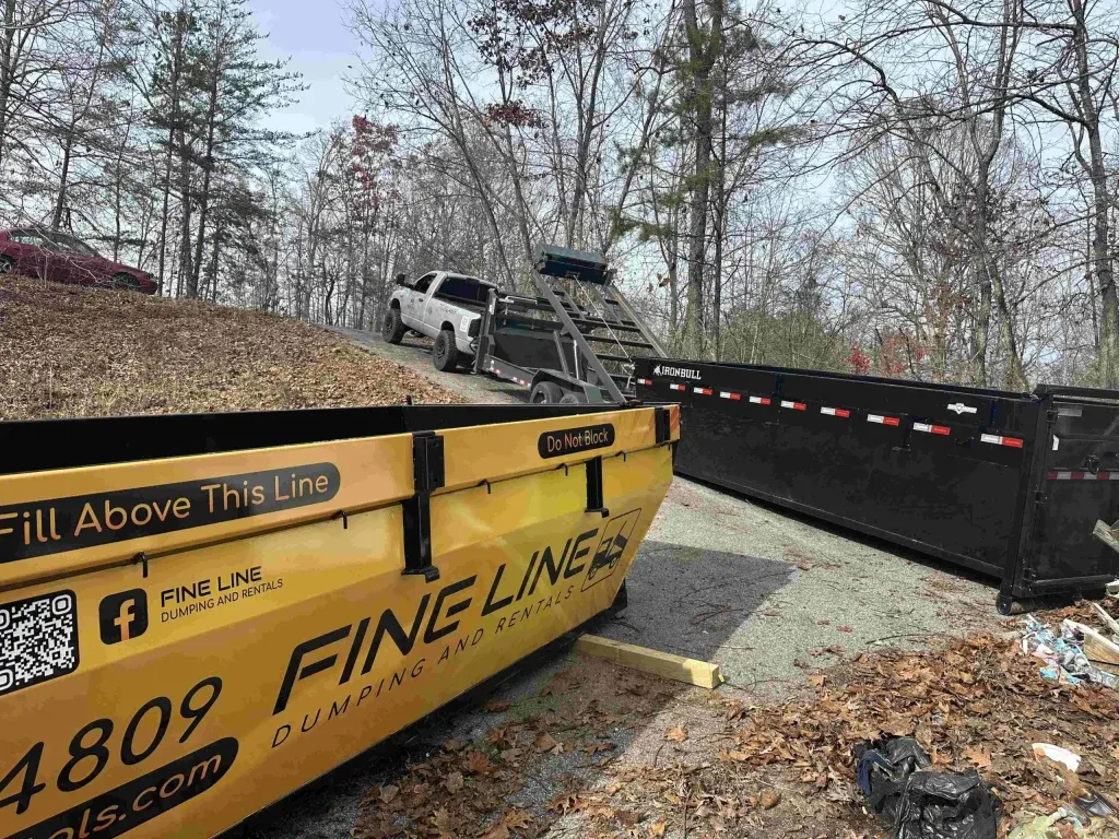 Construction dumpster and debris beside a parked tow truck and black barrier near a wooded roadside.