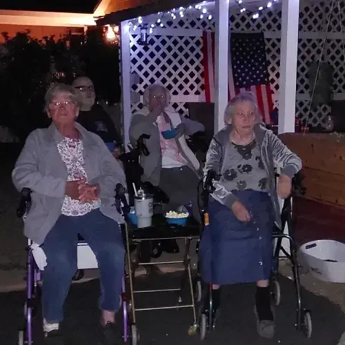 Four elderly people in walkers sit under a decorated porch, an American flag hangs behind them.