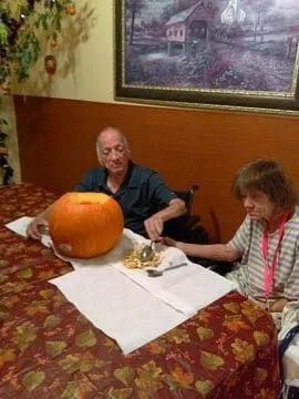 An elderly couple carves a pumpkin at a table. The man sits in a wheelchair, and the woman holds a carving tool.