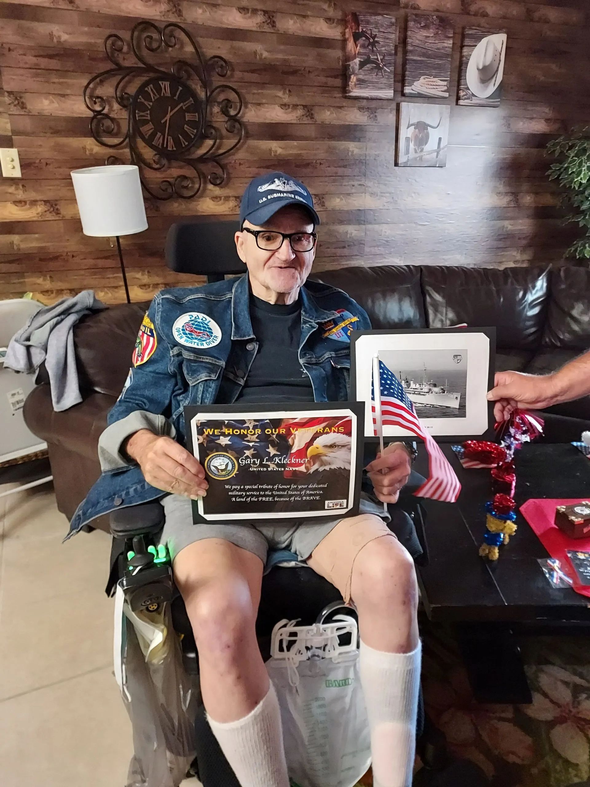 Elderly man in a wheelchair wearing a decorated denim jacket, holding a plaque and small American flag.