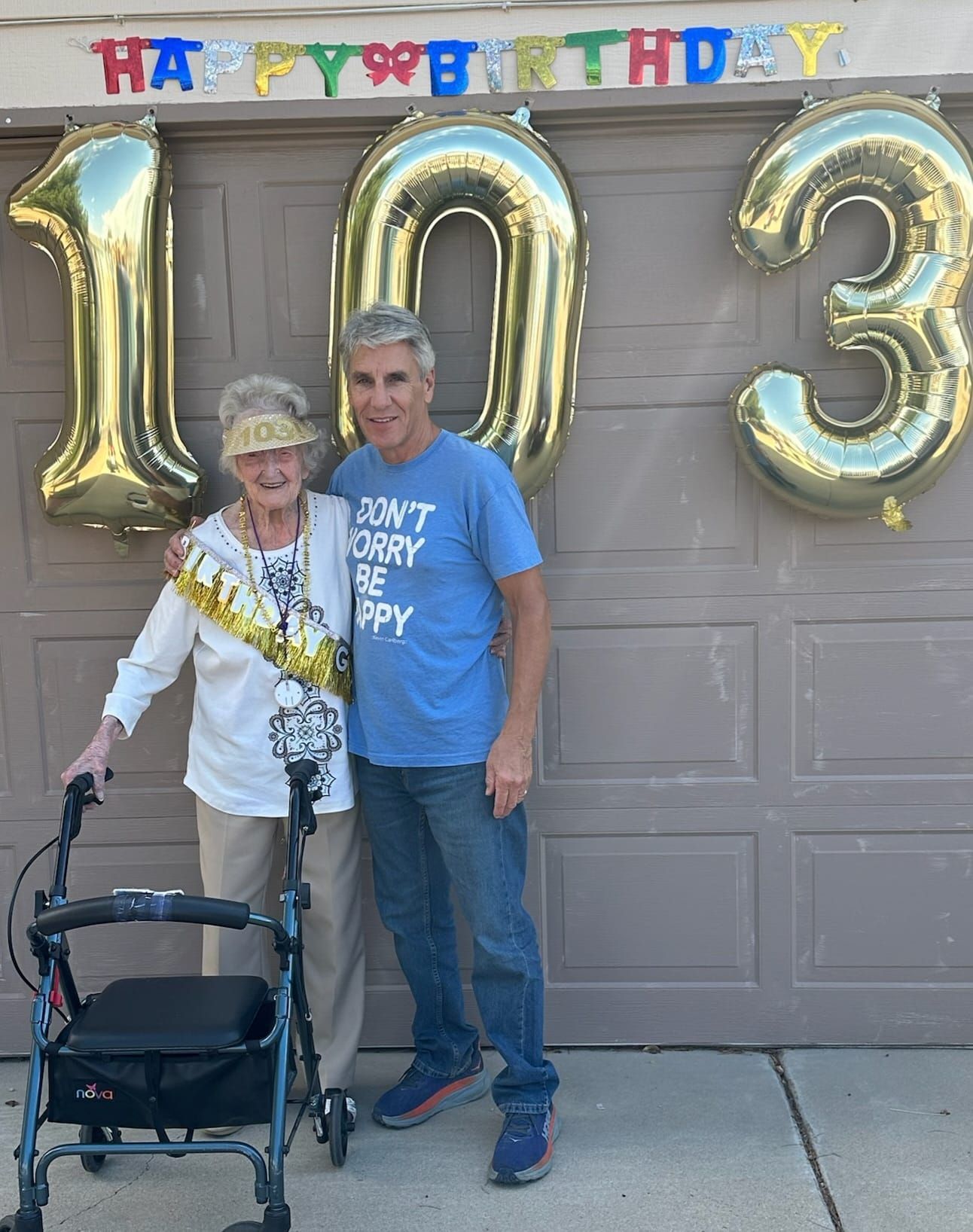 Woman with a gold birthday sash and walker, and a man, pose in front of gold balloons that say 