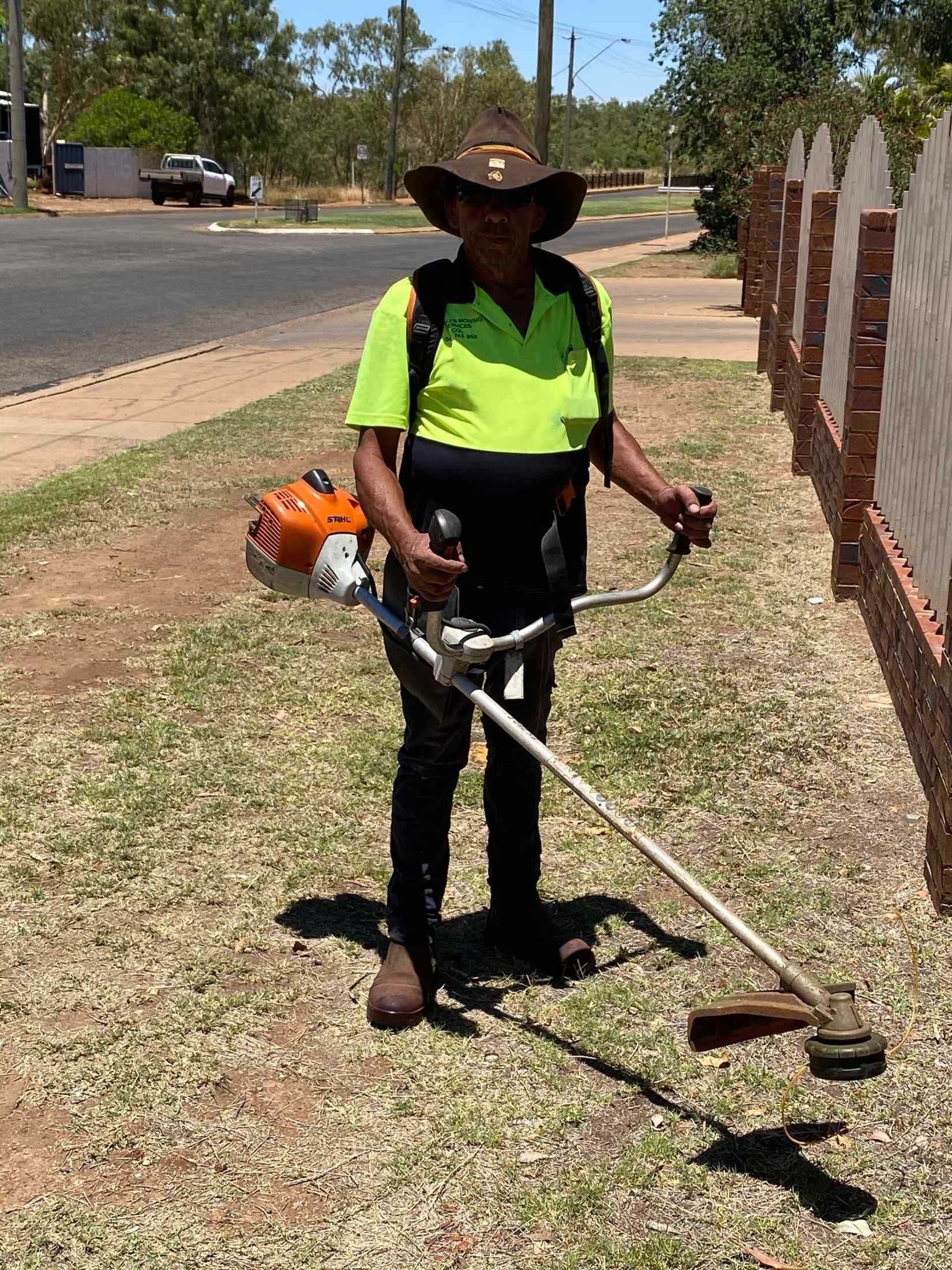 Person Using a Weed Wacker on a Grassy Verge Beside a Street — Darrell's Mowing Services in Sunset, QLD