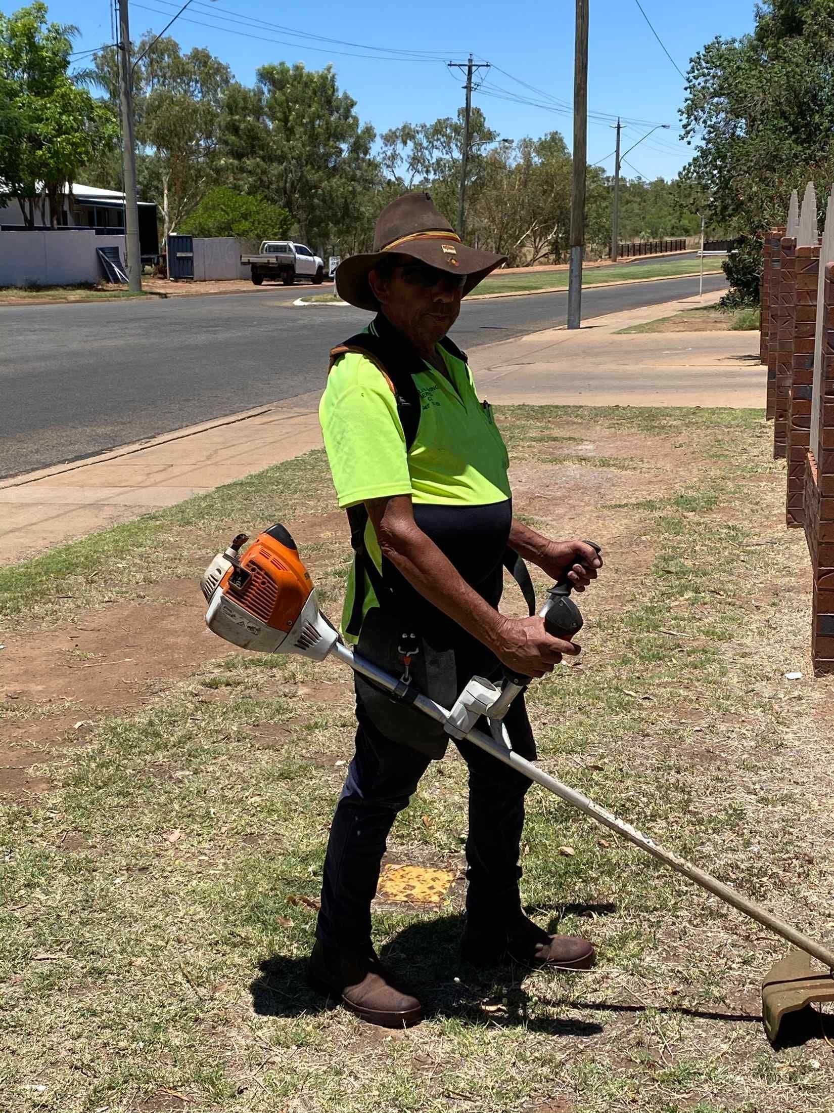 Person Wearing a Hat and Safety Vest Using a Weed Whacker on a Grassy Verge — Darrell's Mowing Services in Sunset, QLD