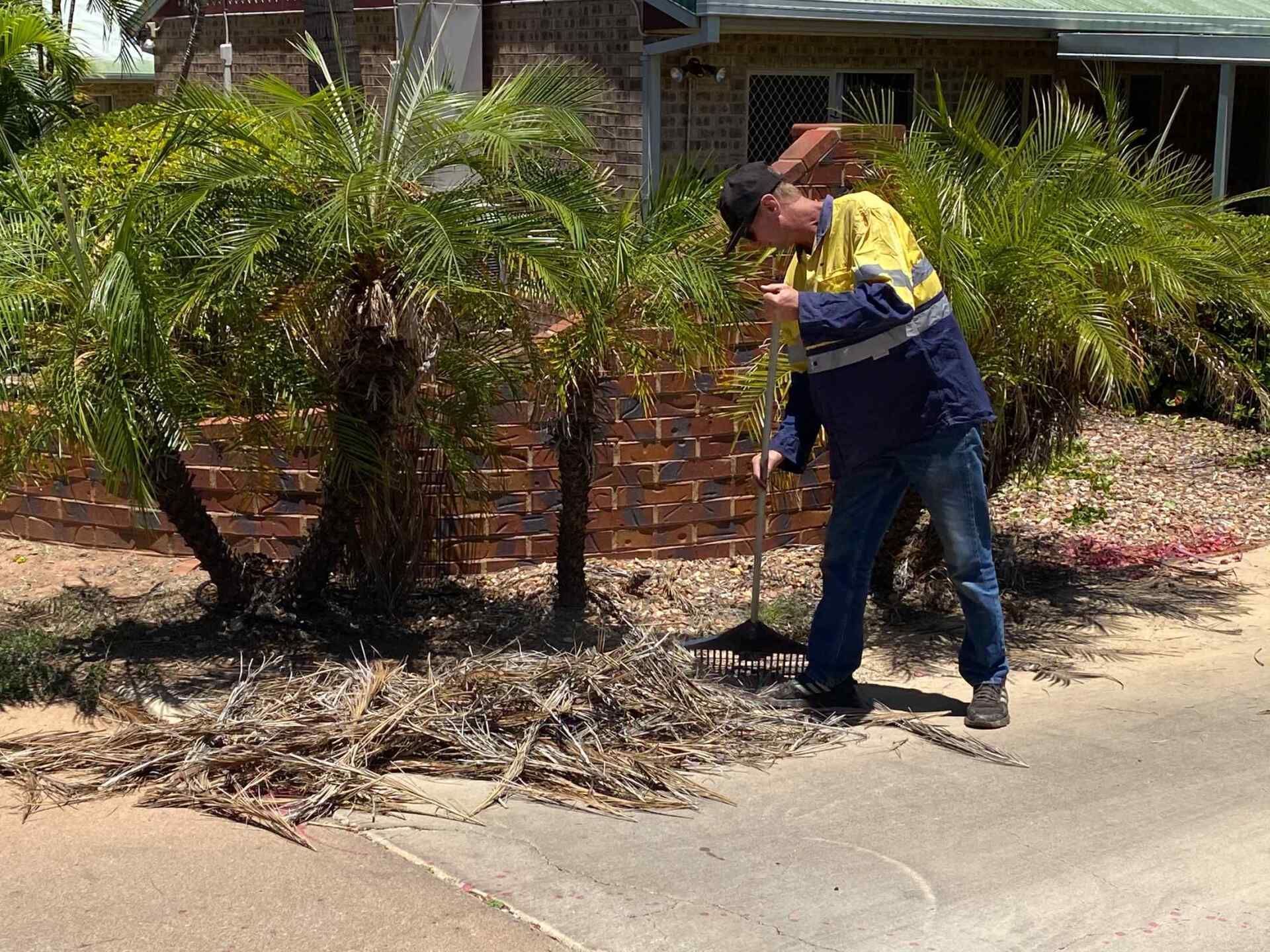 Man Sweeping Debris Near Palm Trees and a Brick Wall — Darrell's Mowing Services in Sunset, QLD