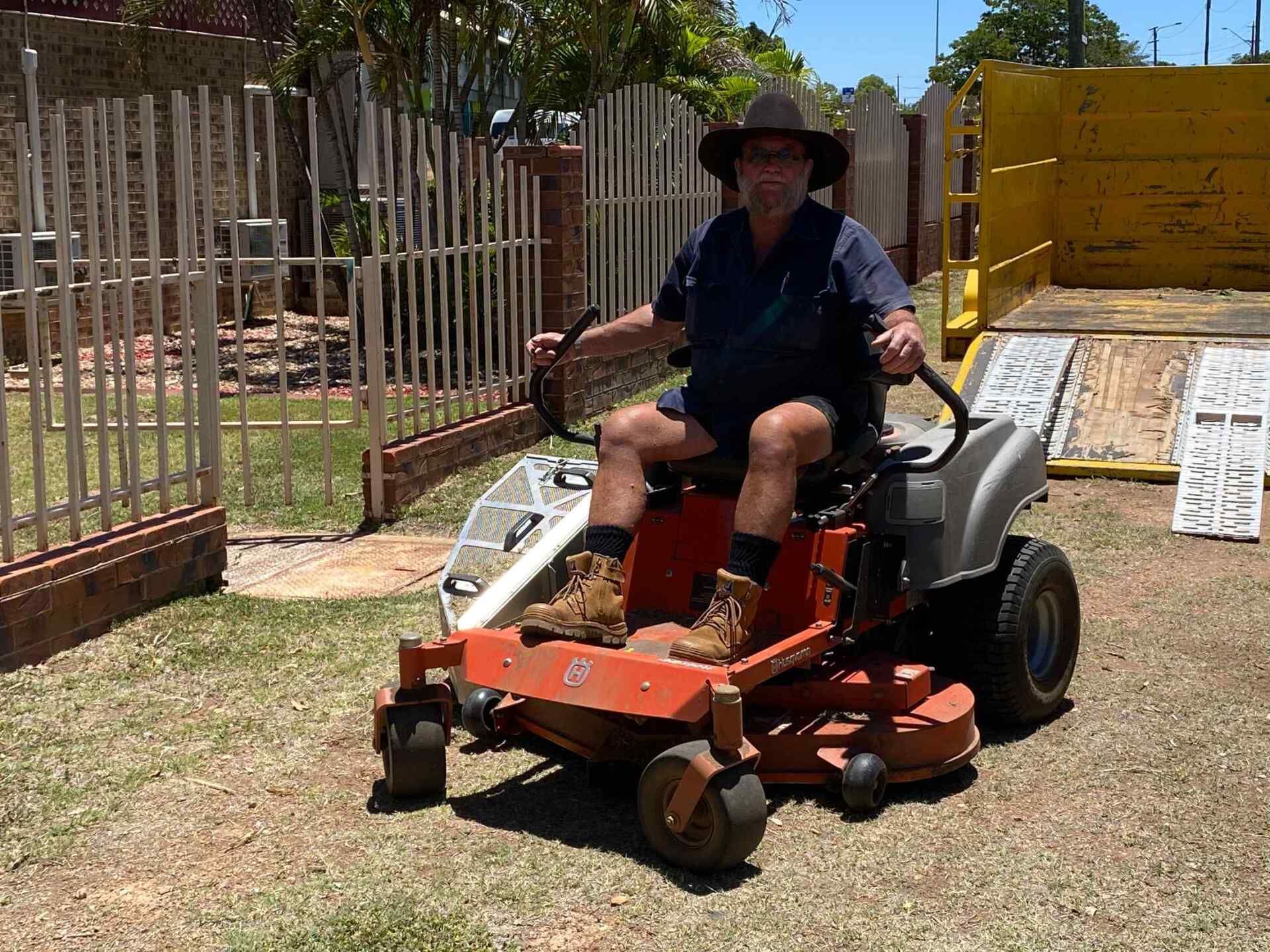 Man on Orange Riding Mower in Front Yard. Brown Hat, Blue Shirt, Shorts, Boots — Darrell's Mowing Services in Sunset, QLD
