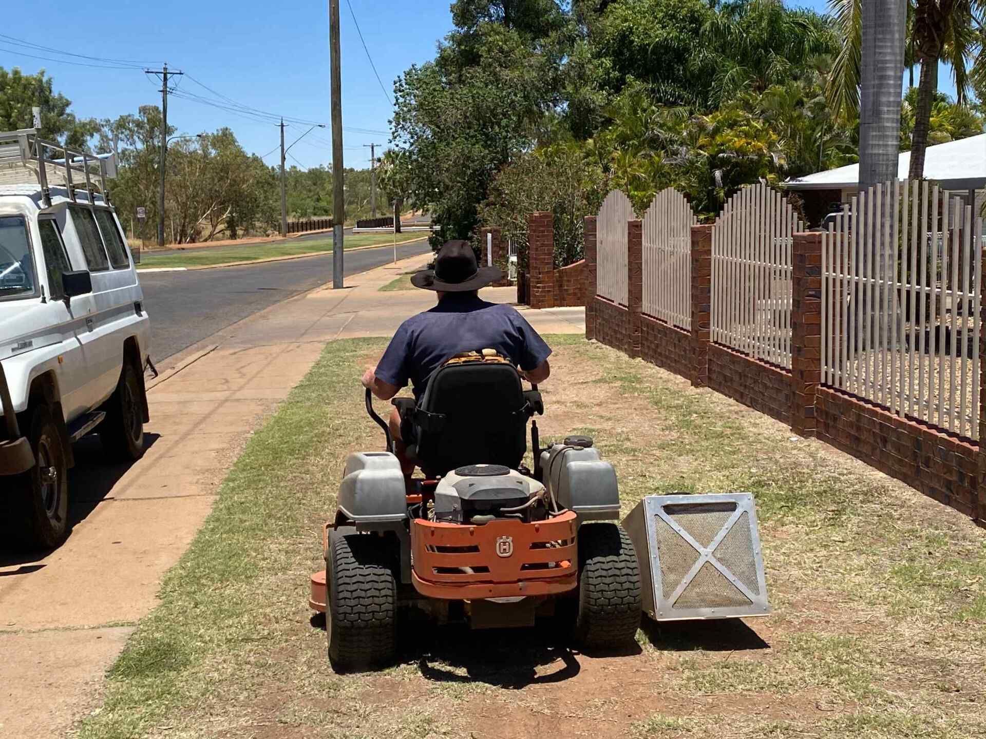 Man on a Riding Lawnmower Mowing Grass Along a Sidewalk Next to a Brick Fence — Darrell's Mowing Services in Sunset, QLD