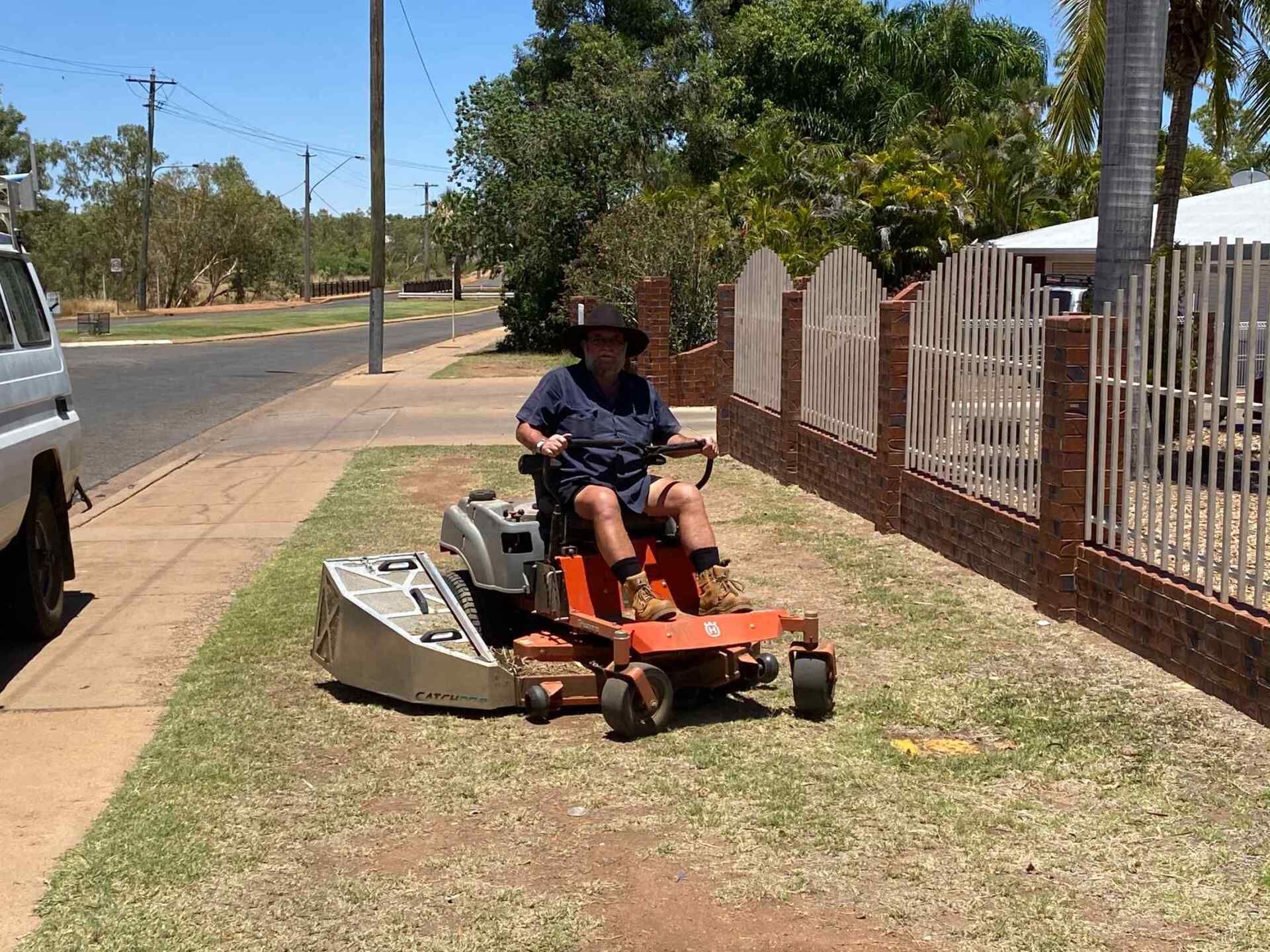 Man Operating a Riding Lawnmower on a Grassy Area Next to a Sidewalk — Darrell's Mowing Services in Sunset, QLD