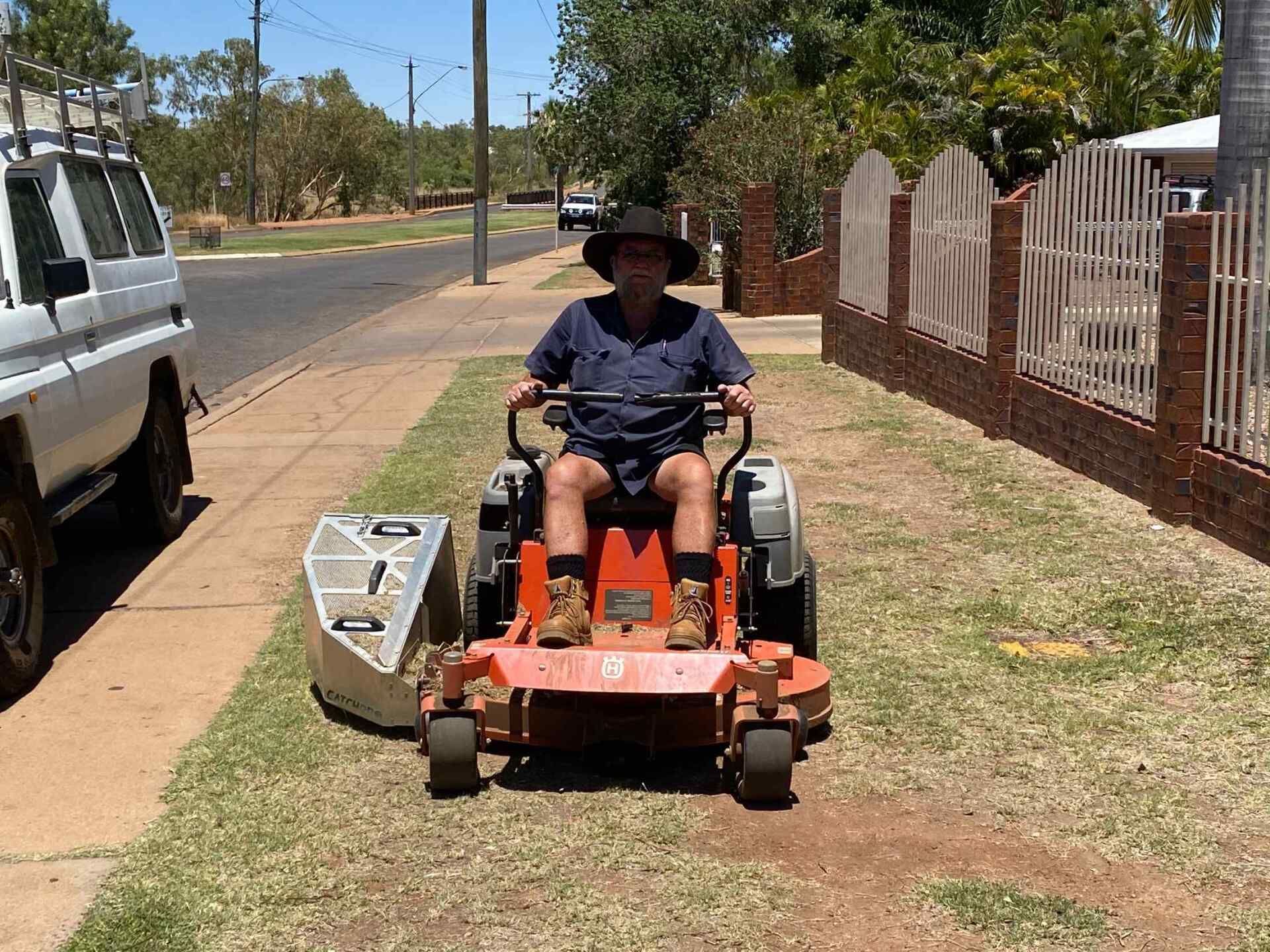Man on a Riding Lawn Mower, Mowing Grass Next to a Sidewalk and a Brick Fence — Darrell's Mowing Services in Sunset, QLD