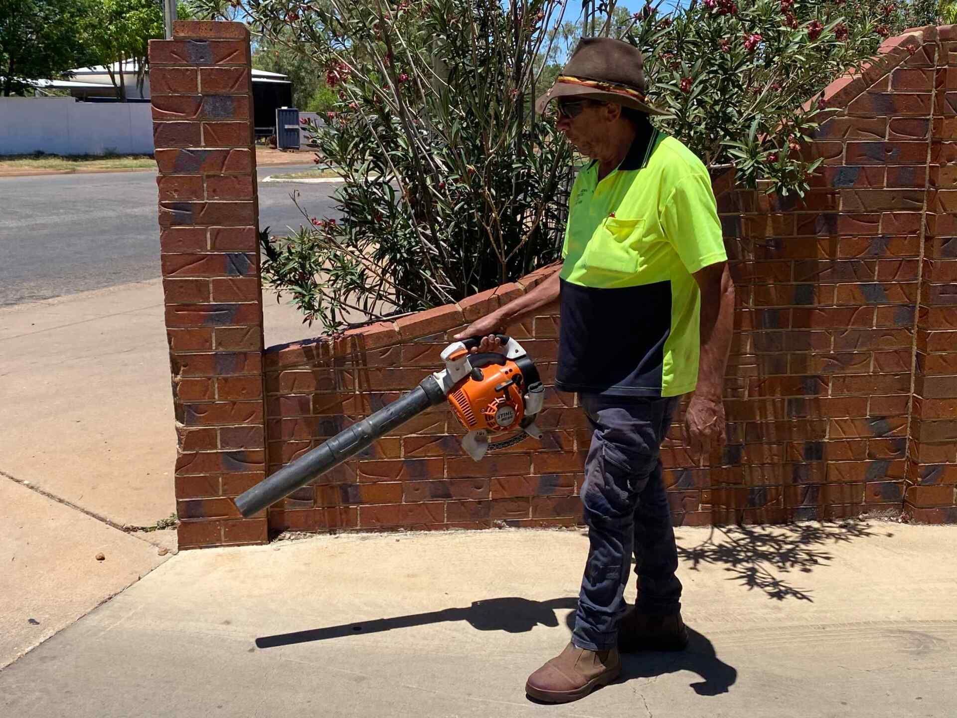 Man in Safety Vest Blowing Debris With a Leaf Blower on a Sidewalk — Darrell's Mowing Services in Sunset, QLD