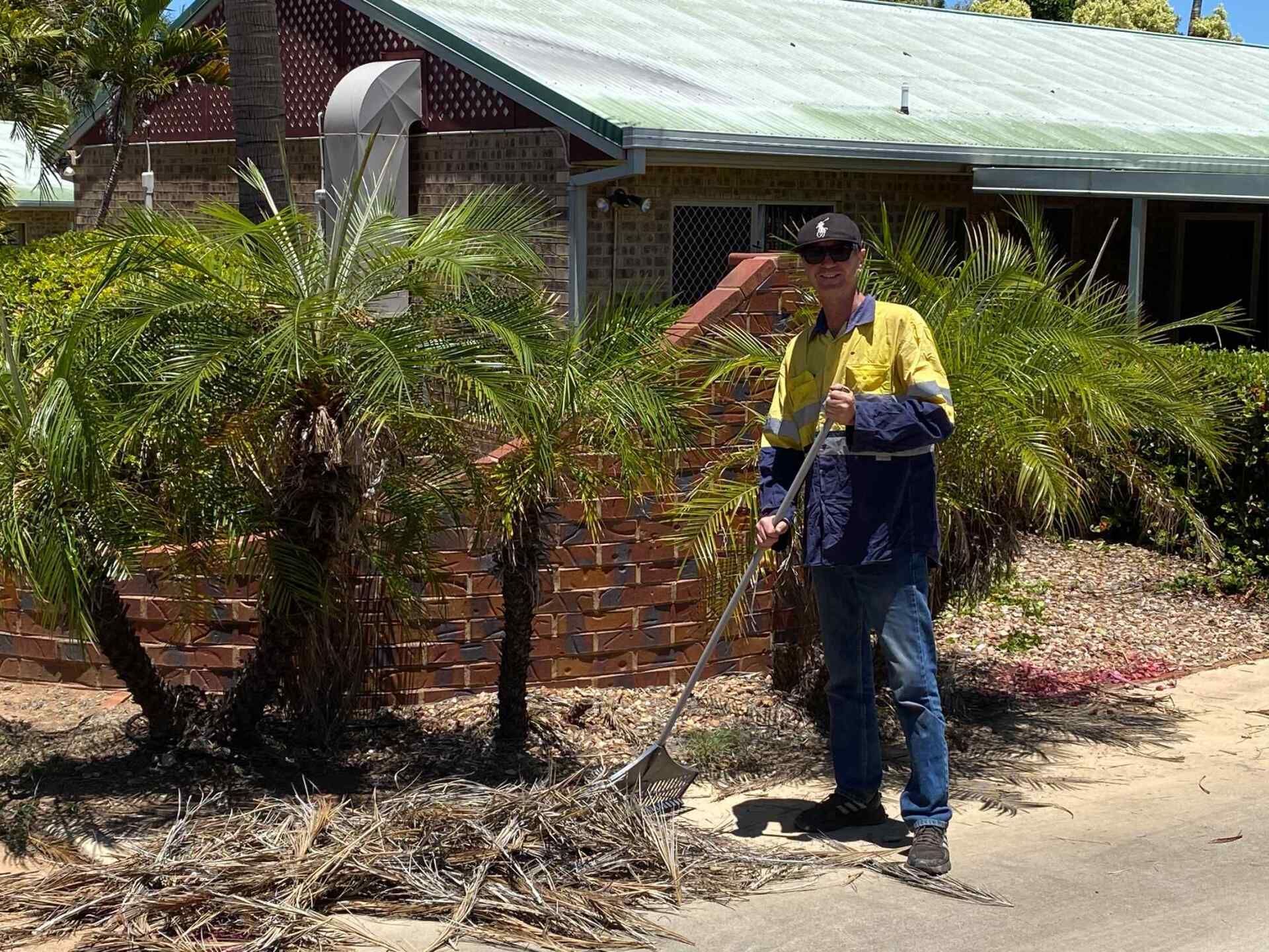 Man in Work Clothes Raking Yard Debris in Front of a Brick House With Palm Trees — Darrell's Mowing Services in Sunset, QLD